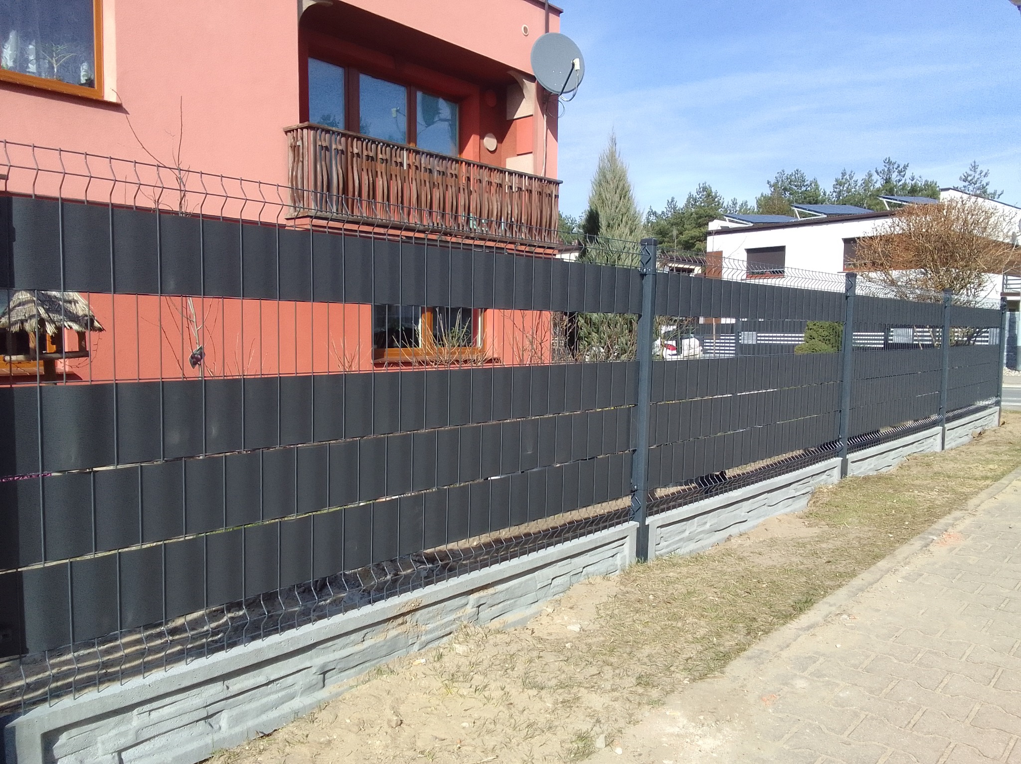 Nowoczesne ogrodzenie panelowe z czarnymi wypełnieniami na betonowej podmurówce, na tle różowego domu z balkonem i zielenią