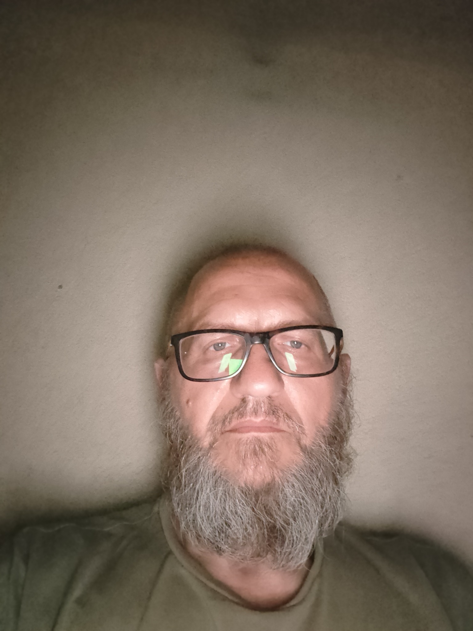 Portret mężczyzny z brodą i okularami na tle jasnej ściany, oświetlonego od góry, tworzącego cień na ścianie.