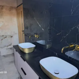 Nowoczesna łazienka w Tczewie z dwoma umywalkami nablatowymi w kolorze białym ze złotym obramowaniem, zamontowanymi na szafce z szufladami w odcieniach szarości, oraz złotą armaturą na tle czarnych...