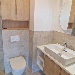 Krzysztof Lica - Nowoczesna łazienka z wiszącą toaletą, umywalką nablatową na szafce z drewnianymi frontami i okrągłym lustrem, wykończona płytkami imitującymi beton w odcieniach szarości.