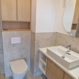 Nowoczesna łazienka z wiszącą toaletą, umywalką nablatową na szafce z drewnianymi frontami i okrągłym lustrem, wykończona płytkami imitującymi beton w odcieniach szarości.