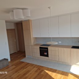 Krzysztof Lica - Jasna kuchnia z drewnianymi szafkami i białymi frontami górnymi, czarny zlewozmywak i płyta grzewcza, drewniana podłoga i szare płytki pod szafkami, minimalistyczne oświetlenie.