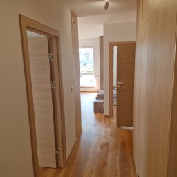 Krzysztof Lica - Korytarz w mieszkaniu z drewnianą podłogą, dwiema parami drzwi z drewnianymi ościeżnicami i zabudowaną szafą z jasnego drewna, oświetlony reflektorami sufitowymi.