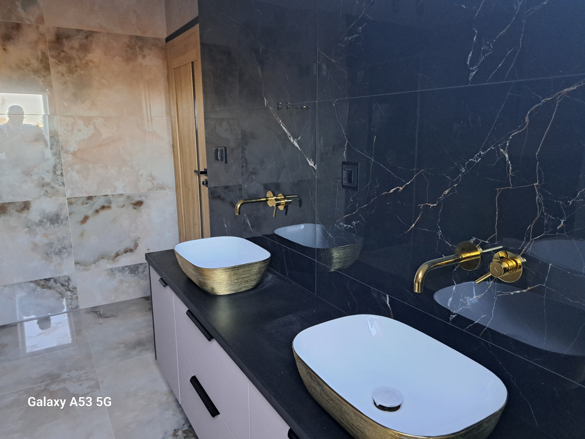 Nowoczesna łazienka w Tczewie z dwoma umywalkami nablatowymi w kolorze białym ze złotym obramowaniem, zamontowanymi na szafce z szufladami w odcieniach szarości, oraz złotą armaturą na tle czarnych...