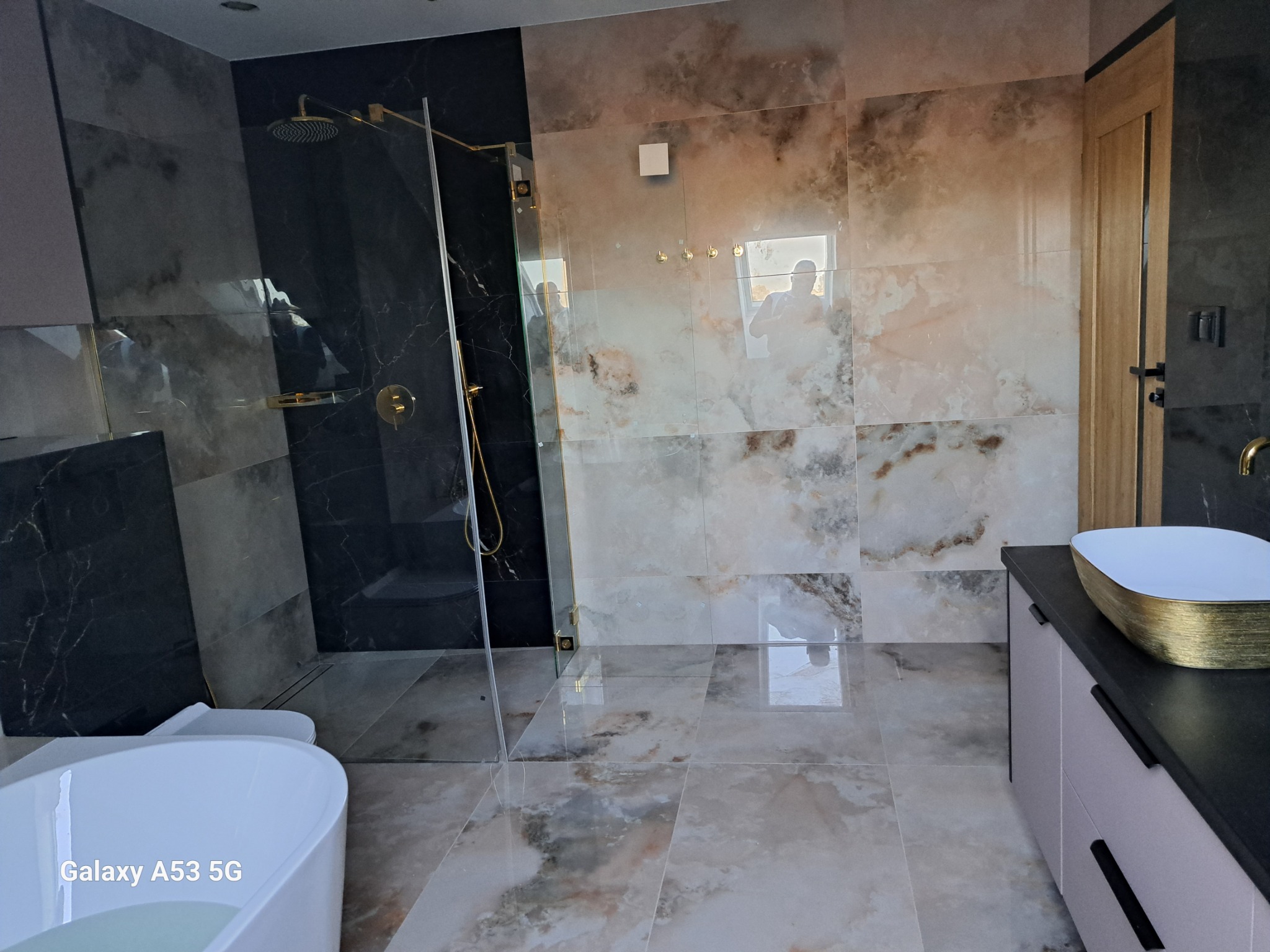 Nowoczesna łazienka z kabiną prysznicową typu walk-in, umywalką nablatową ze złotymi akcentami i płytkami imitującymi marmur na ścianach i podłodze. Widoczna wanna wolnostojąca z białym wykończeniem.