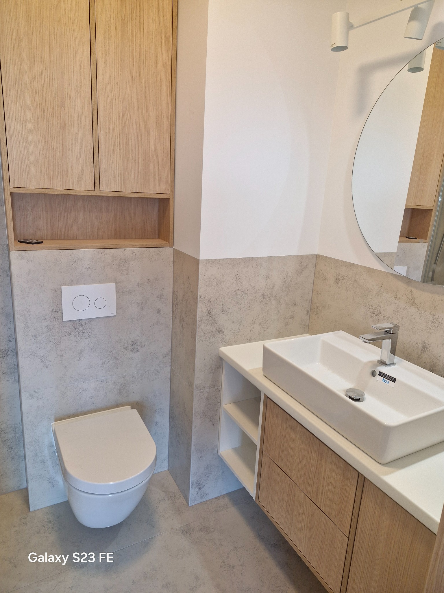 Nowoczesna łazienka z wiszącą toaletą, umywalką nablatową na szafce z drewnianymi frontami i okrągłym lustrem, wykończona płytkami imitującymi beton w odcieniach szarości.