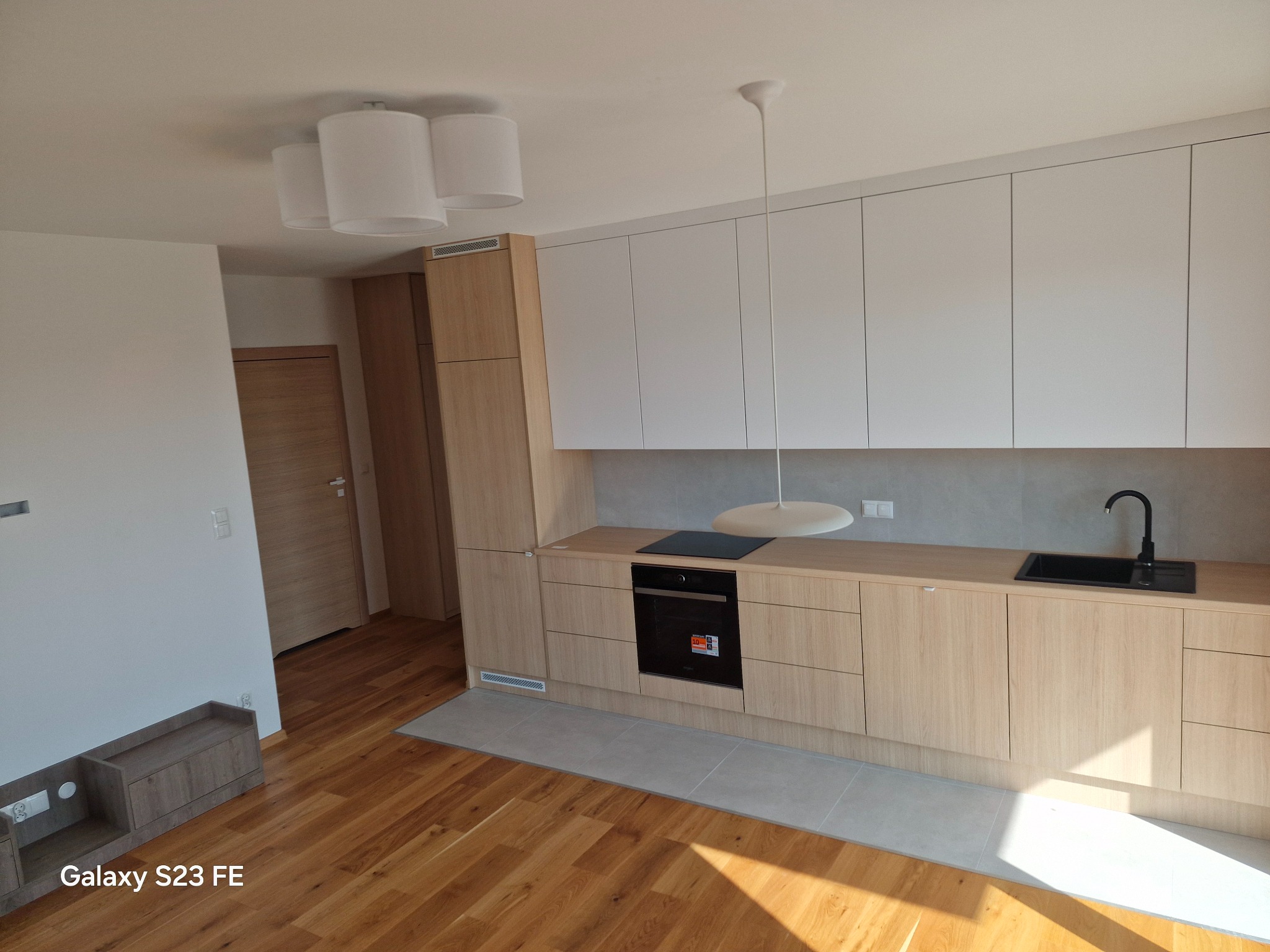 Jasna kuchnia z drewnianymi szafkami i białymi frontami górnymi, czarny zlewozmywak i płyta grzewcza, drewniana podłoga i szare płytki pod szafkami, minimalistyczne oświetlenie.