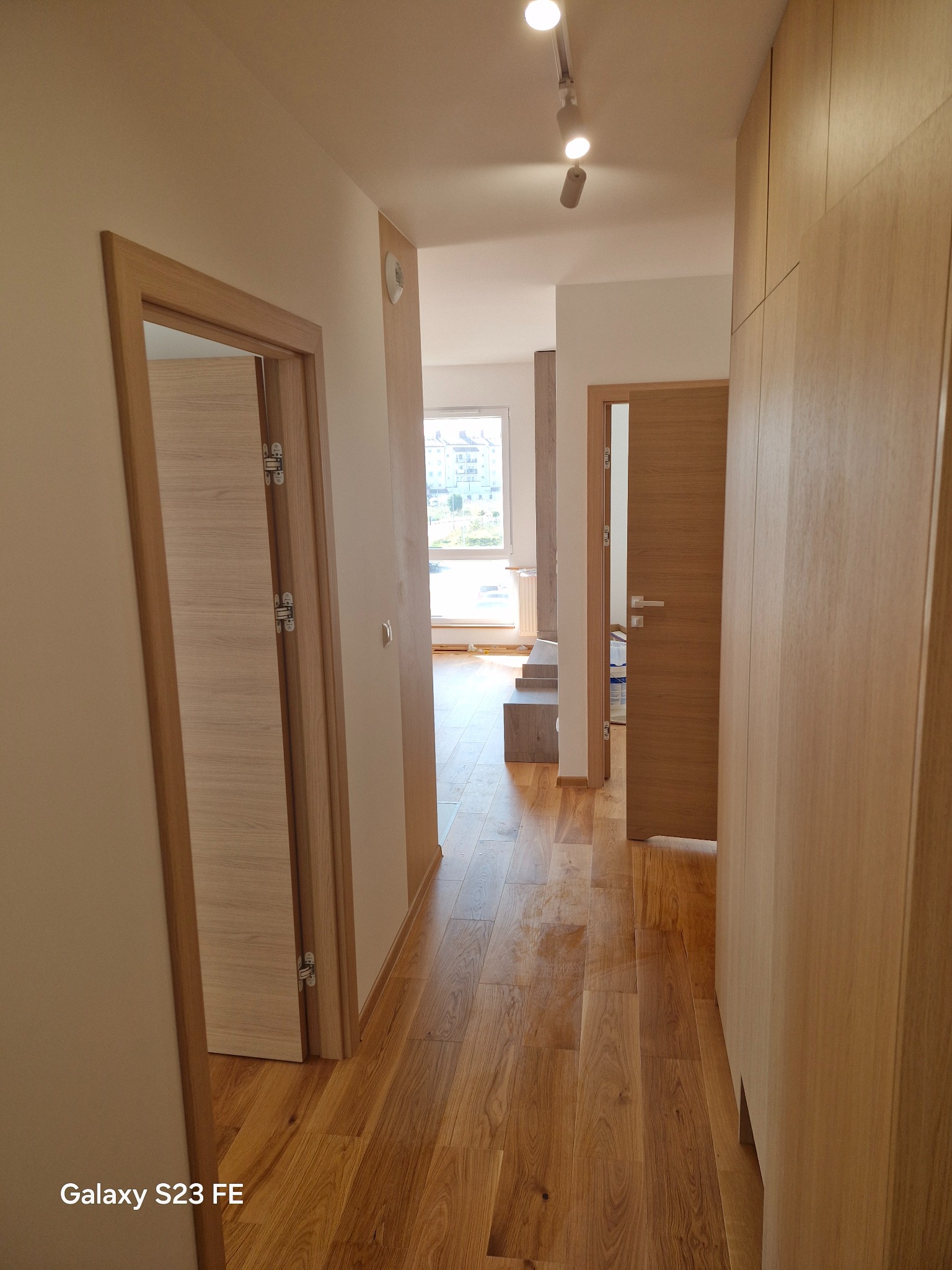 Korytarz w mieszkaniu z drewnianą podłogą, dwiema parami drzwi z drewnianymi ościeżnicami i zabudowaną szafą z jasnego drewna, oświetlony reflektorami sufitowymi.