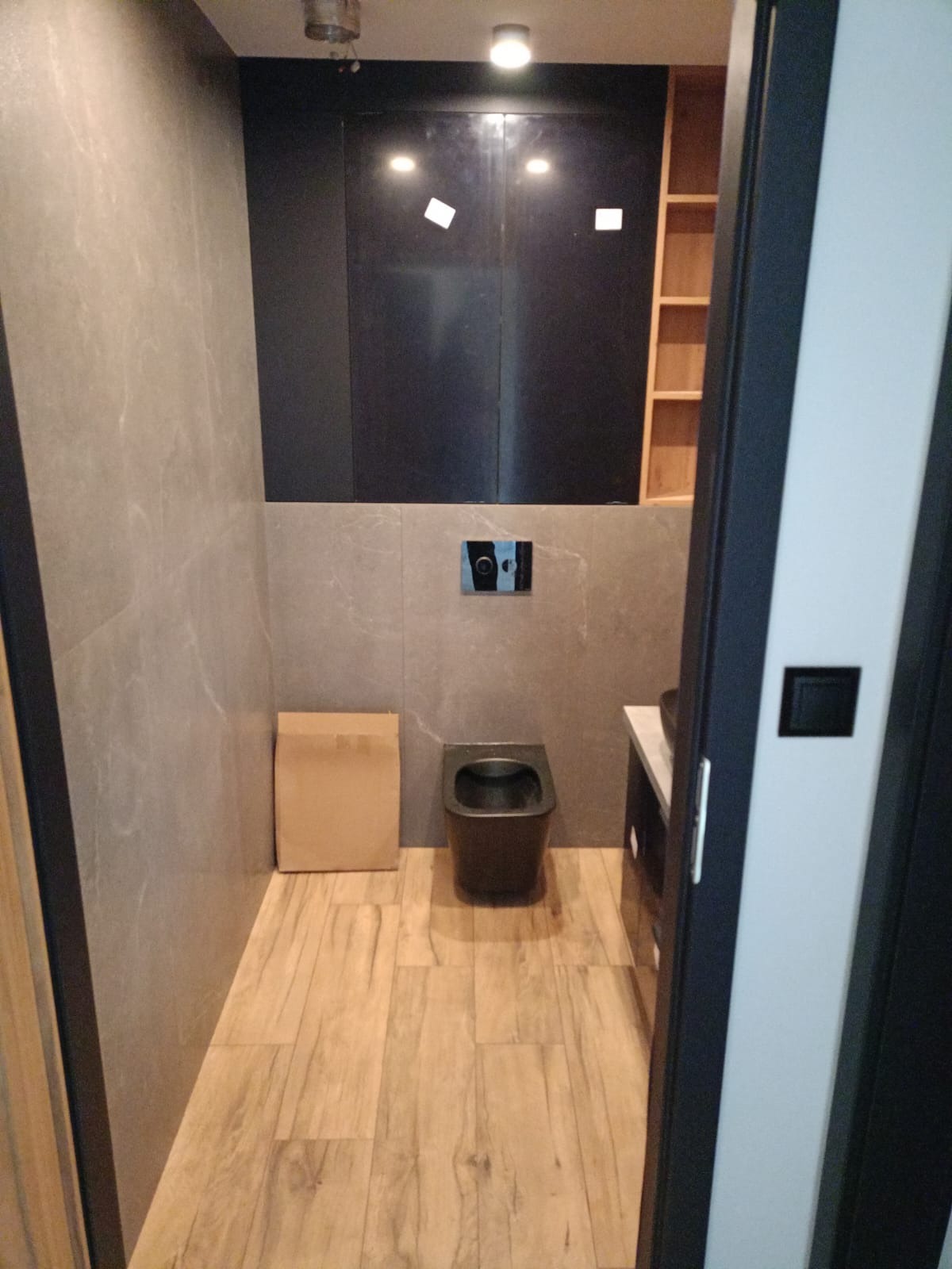 Nowoczesna łazienka z szarą marmurową okładziną ścienną, drewnopodobną podłogą, podwieszaną miską WC w kolorze antracytowym oraz zabudową meblową z czarnymi frontami, widoczna kartonowa paczka.