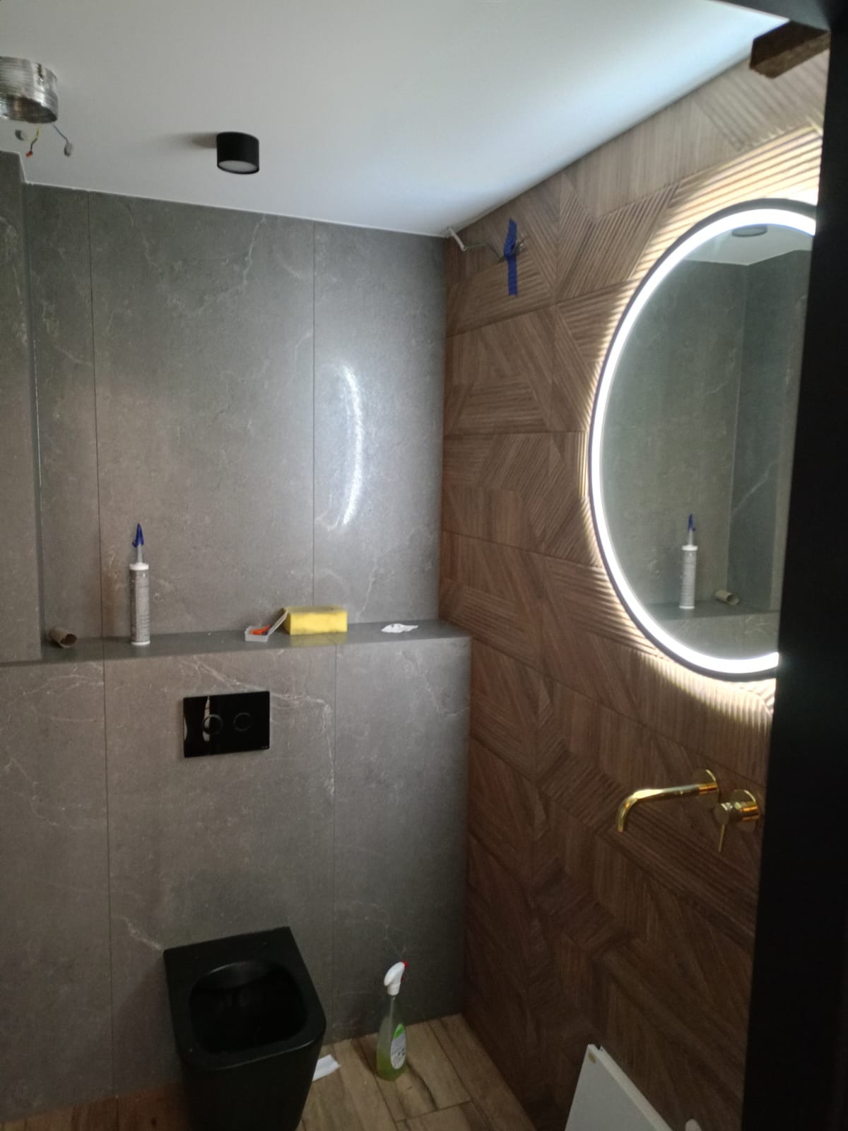 Nowoczesna łazienka w trakcie wykańczania: szare płytki imitujące kamień, drewnopodobne panele ścienne, okrągłe lustro z podświetleniem LED, czarna miska WC i złota bateria umywalkowa, widoczne...