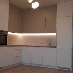 Rad Meble Piotr Radzioch - Nowoczesna kuchnia na wymiar w odcieniach bieli i szarości z podświetleniem LED pod szafkami wiszącymi, czarna bateria kuchenna i widok na korytarz.