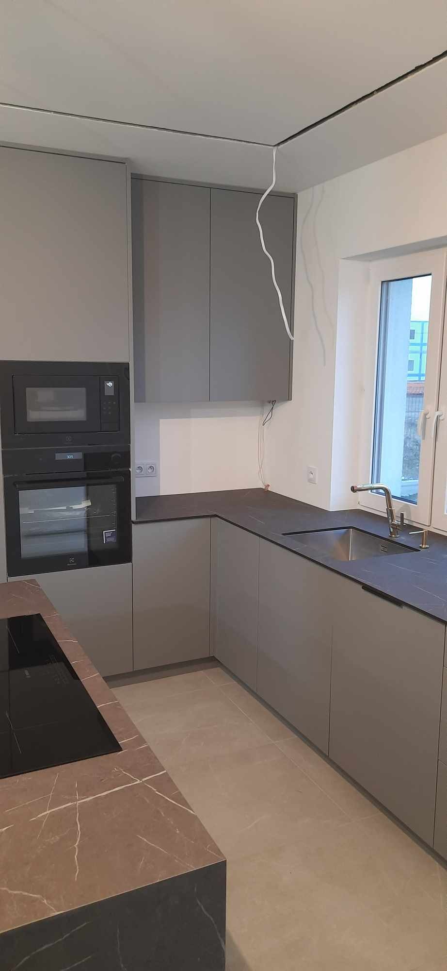 Szara, nowoczesna kuchnia z blatem imitującym marmur, wbudowanym piekarnikiem i mikrofalówką, oraz niedokończonymi kablami wychodzącymi ze ściany.