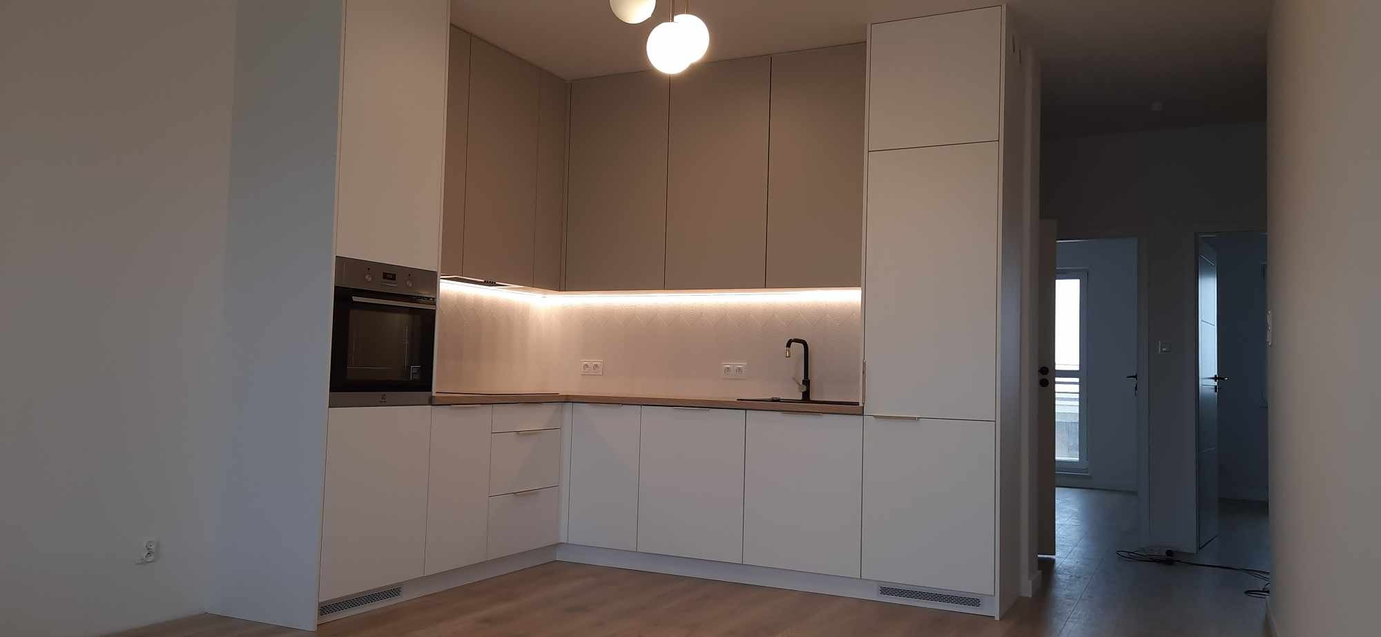 Nowoczesna kuchnia na wymiar w odcieniach bieli i szarości z podświetleniem LED pod szafkami wiszącymi, czarna bateria kuchenna i widok na korytarz.