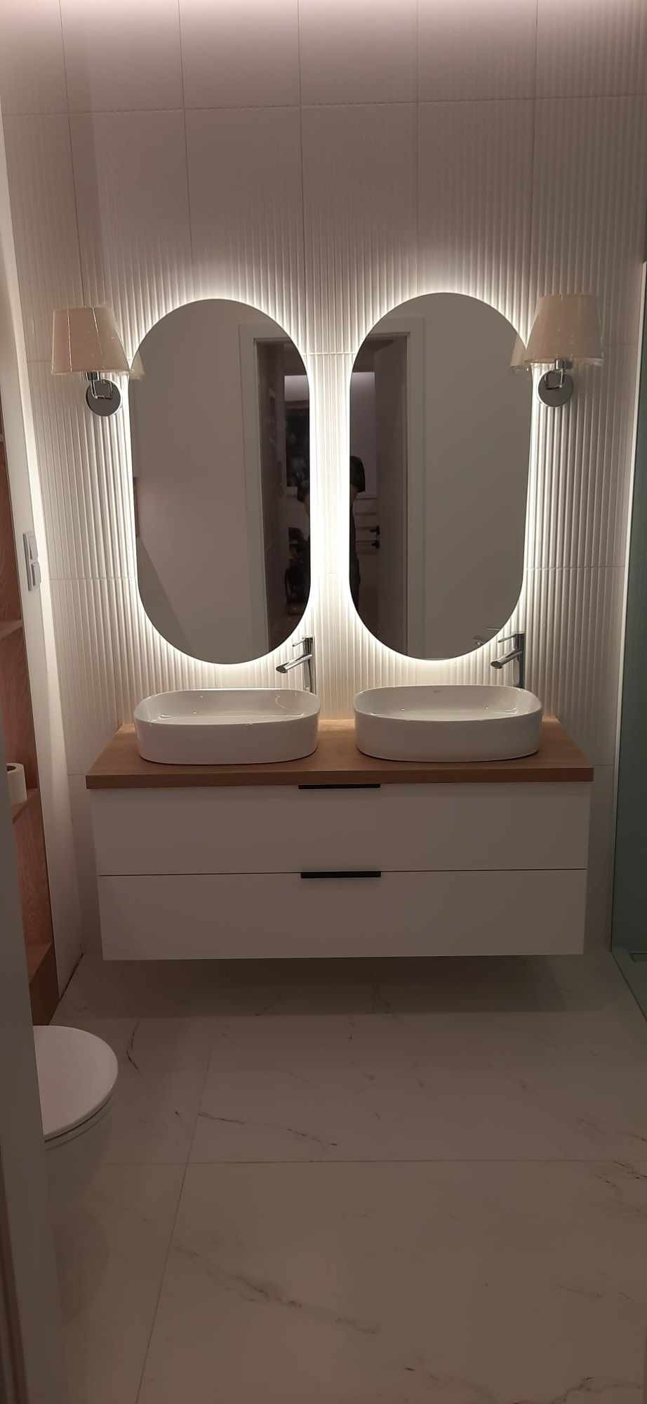 Nowoczesna łazienka w Iławie z podwójną umywalką nablatową na drewnianym blacie i białymi szafkami z minimalistycznymi uchwytami, podświetlane owalne lustra i teksturowane płytki ścienne.