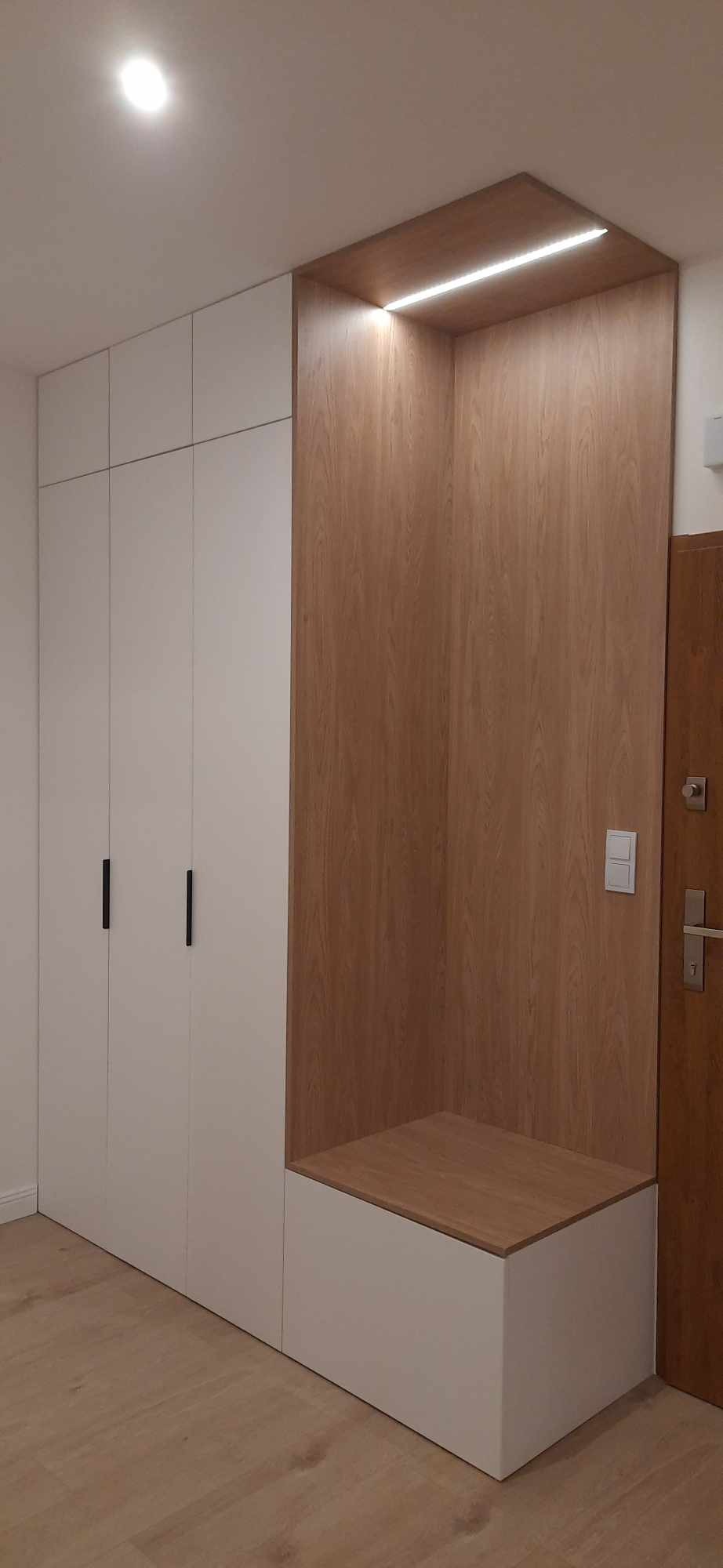 Biała szafa z czarnymi uchwytami i drewniany panel z siedziskiem oraz oświetleniem LED w przedpokoju, widok z dołu.