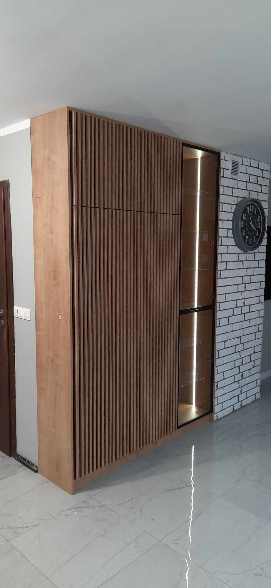 Nowoczesna szafa na wymiar z drewnianymi lamelami i przeszkloną witryną z oświetleniem LED, umieszczona przy ścianie z białej cegły i dużym zegarem.
