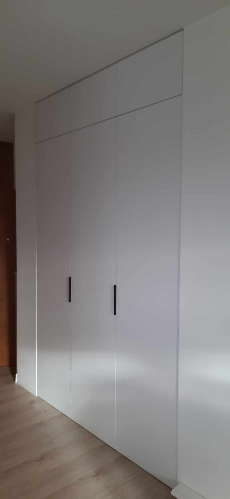 Biała szafa wnękowa z minimalistycznymi, czarnymi uchwytami, zabudowana od podłogi do sufitu, z widocznym fragmentem drewnianej podłogi i drzwi w tle.