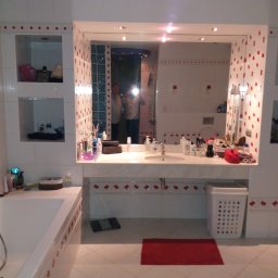 Jerzy Słowiński - Wykończona łazienka w białych płytkach z czerwonymi akcentami kwiatowymi, widoczna wanna, umywalka z marmurowym blatem i lustro, w którym odbija się osoba robiąca zdjęcie.