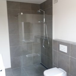 Jerzy Słowiński - Nowoczesna kabina prysznicowa walk-in z szarymi płytkami i podtynkową baterią, widoczny podwieszany sedes z białym przyciskiem spłukującym na tle płytek.