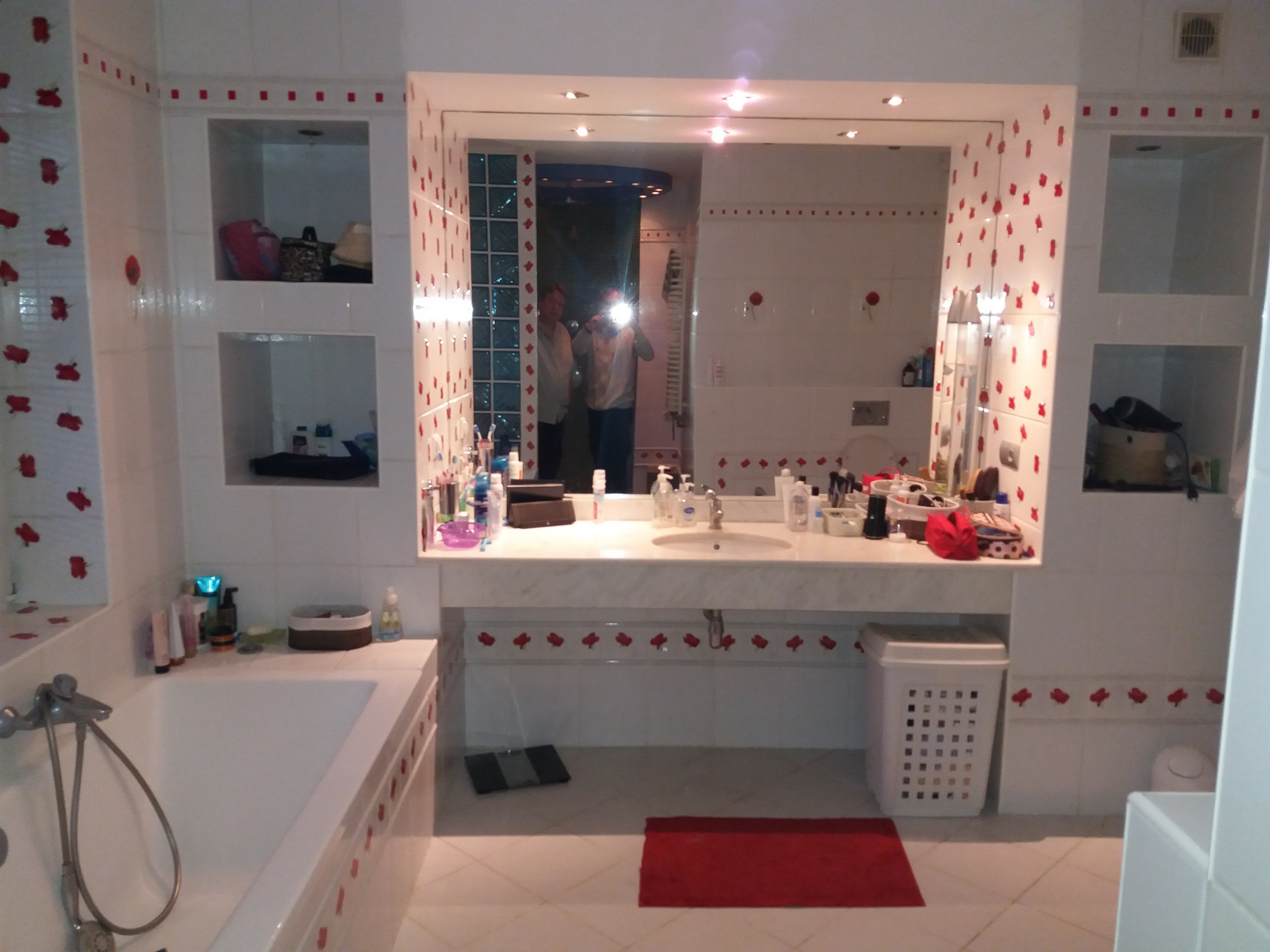 Wykończona łazienka w białych płytkach z czerwonymi akcentami kwiatowymi, widoczna wanna, umywalka z marmurowym blatem i lustro, w którym odbija się osoba robiąca zdjęcie.
