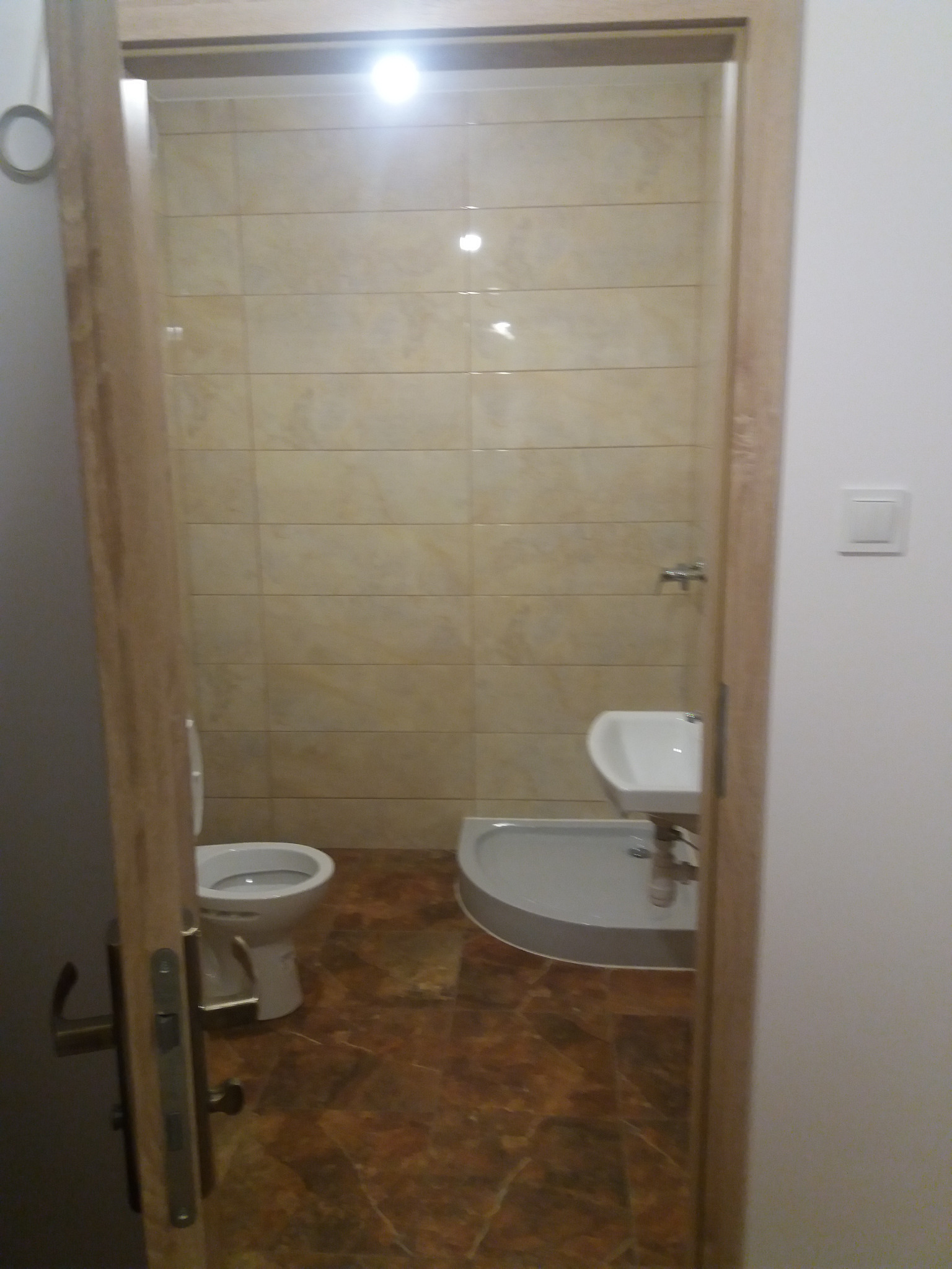 Wykończona łazienka z beżowymi płytkami i brązową podłogą, widok z korytarza w Legionowie. Widoczna toaleta, umywalka i kabina prysznicowa.