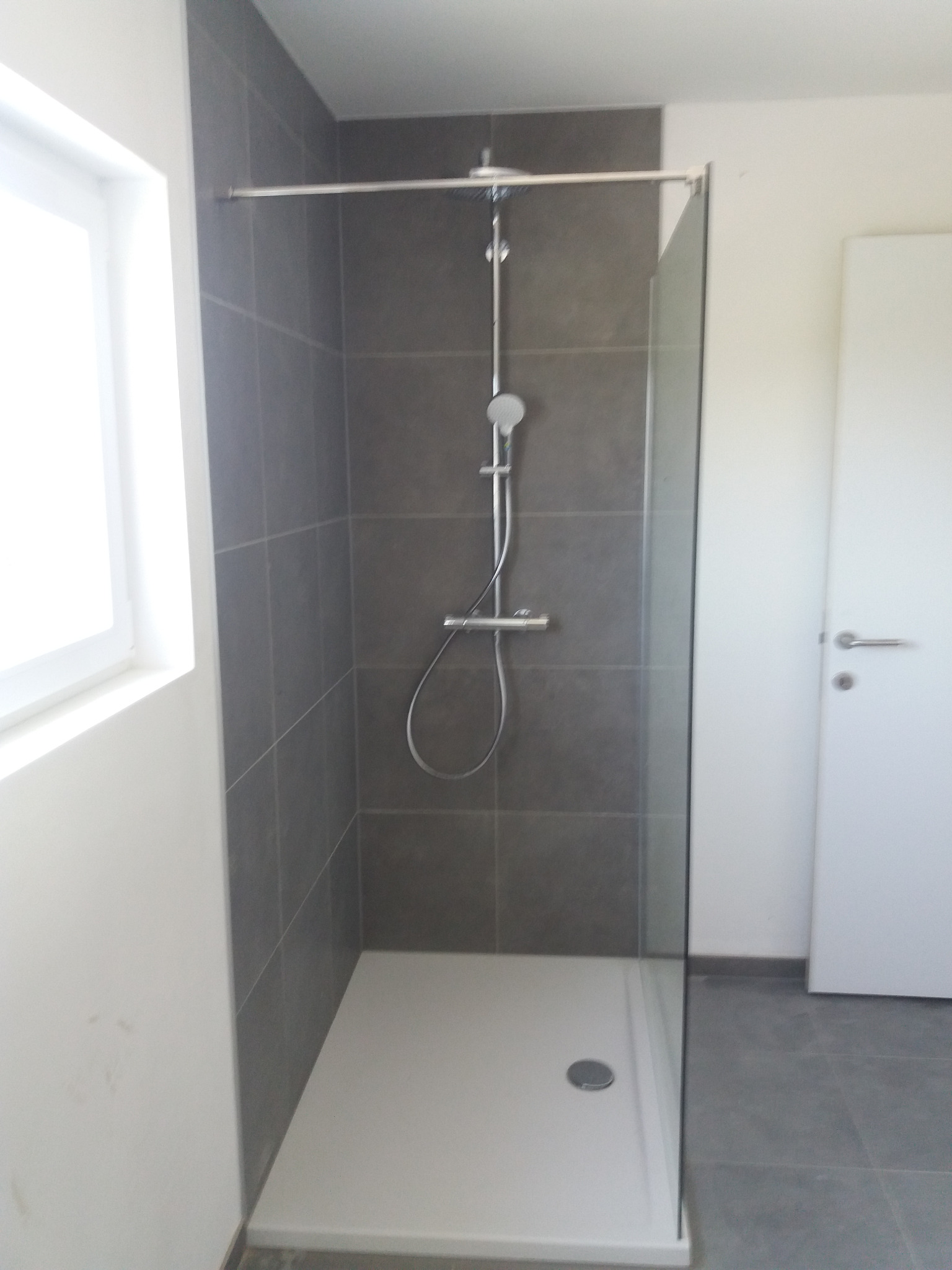 Nowoczesna kabina prysznicowa z szarymi płytkami na ścianach, biały brodzik i szklana szyba, widok na białe drzwi w tle.