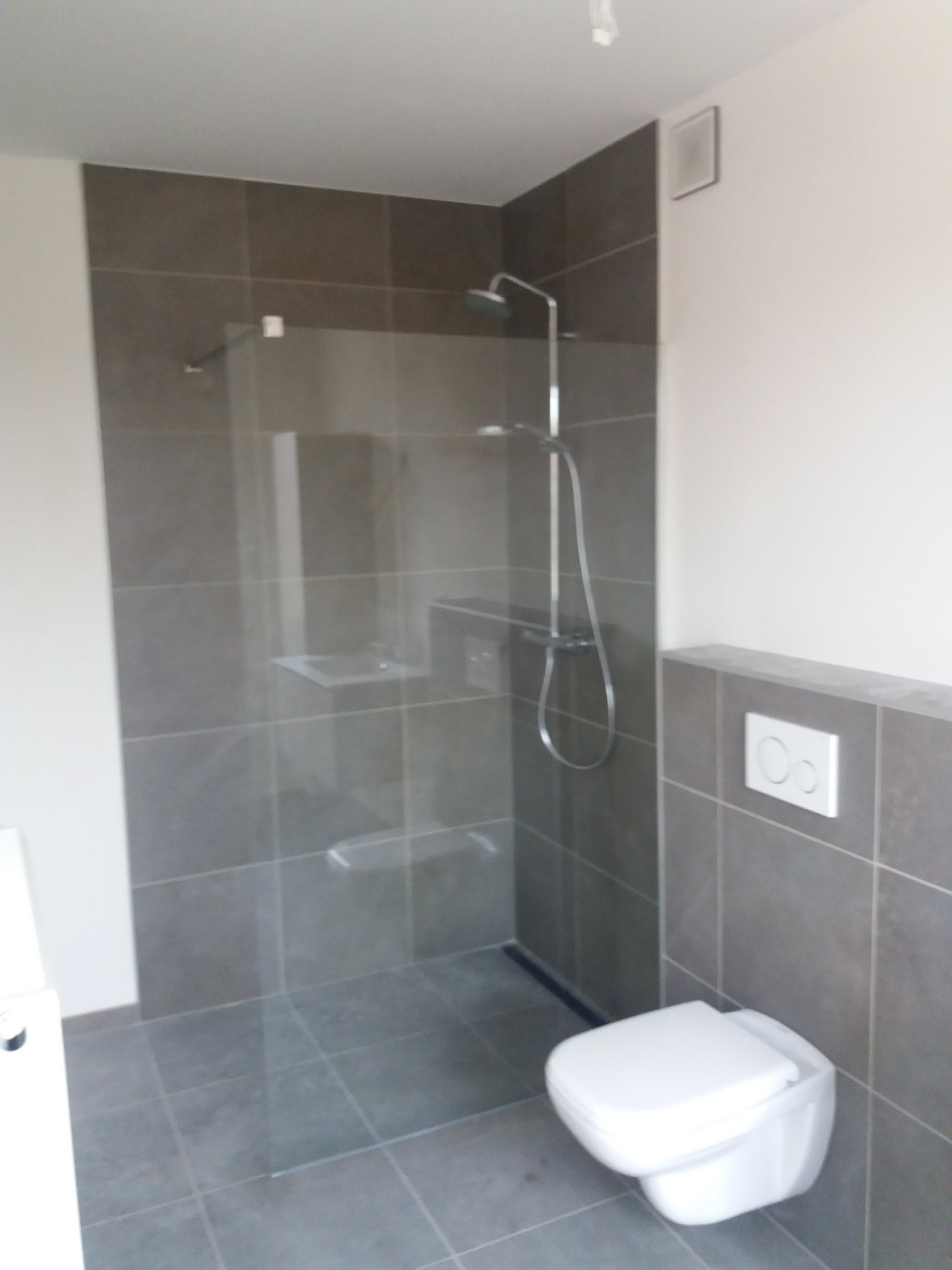 Nowoczesna kabina prysznicowa walk-in z szarymi płytkami i podtynkową baterią, widoczny podwieszany sedes z białym przyciskiem spłukującym na tle płytek.