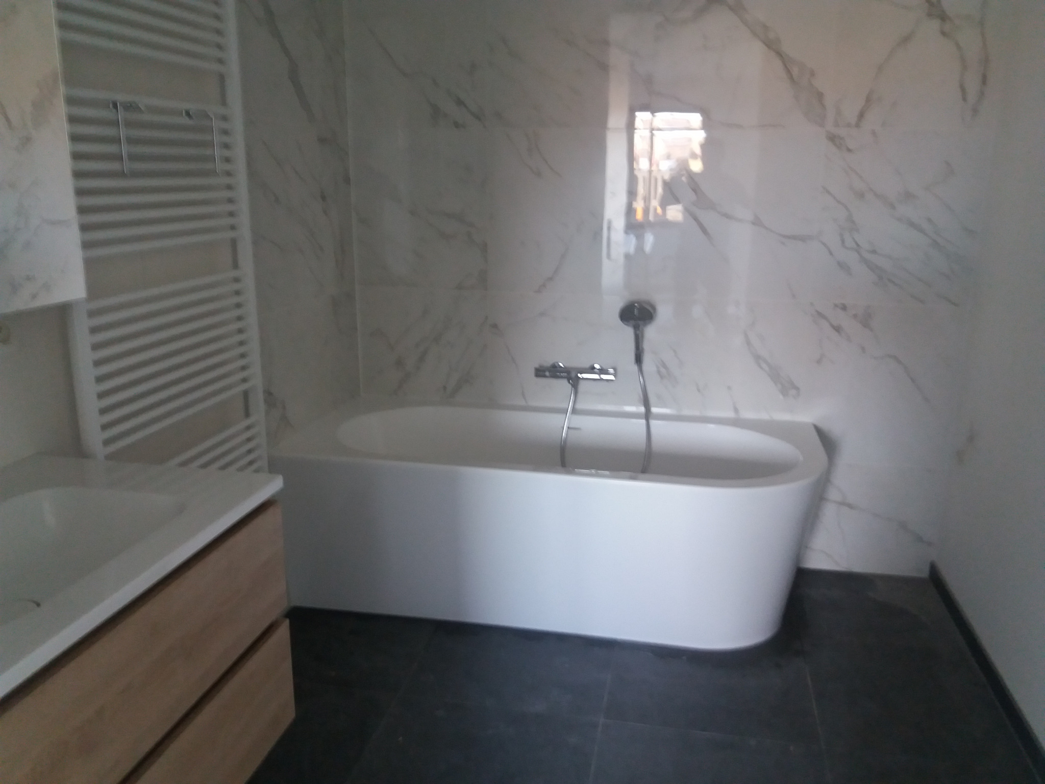 Nowoczesna łazienka z białą, wolnostojącą wanną, grzejnikiem drabinkowym, umywalką z drewnianą szafką i marmurowymi płytkami na ścianach.