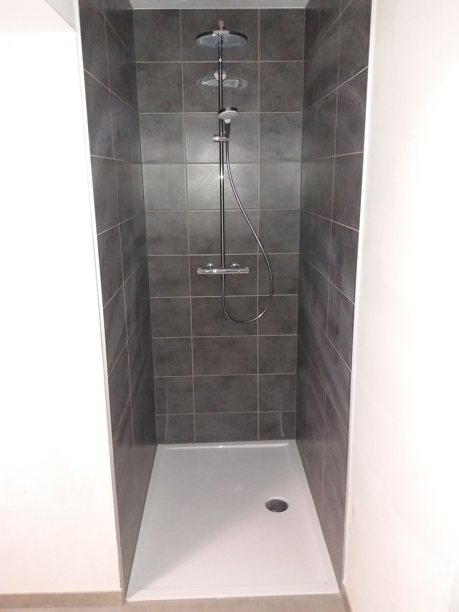 Wnętrze kabiny prysznicowej wyłożonej ciemnoszarą mozaiką, z deszczownicą i baterią w kolorze chromu oraz białym brodzikiem.