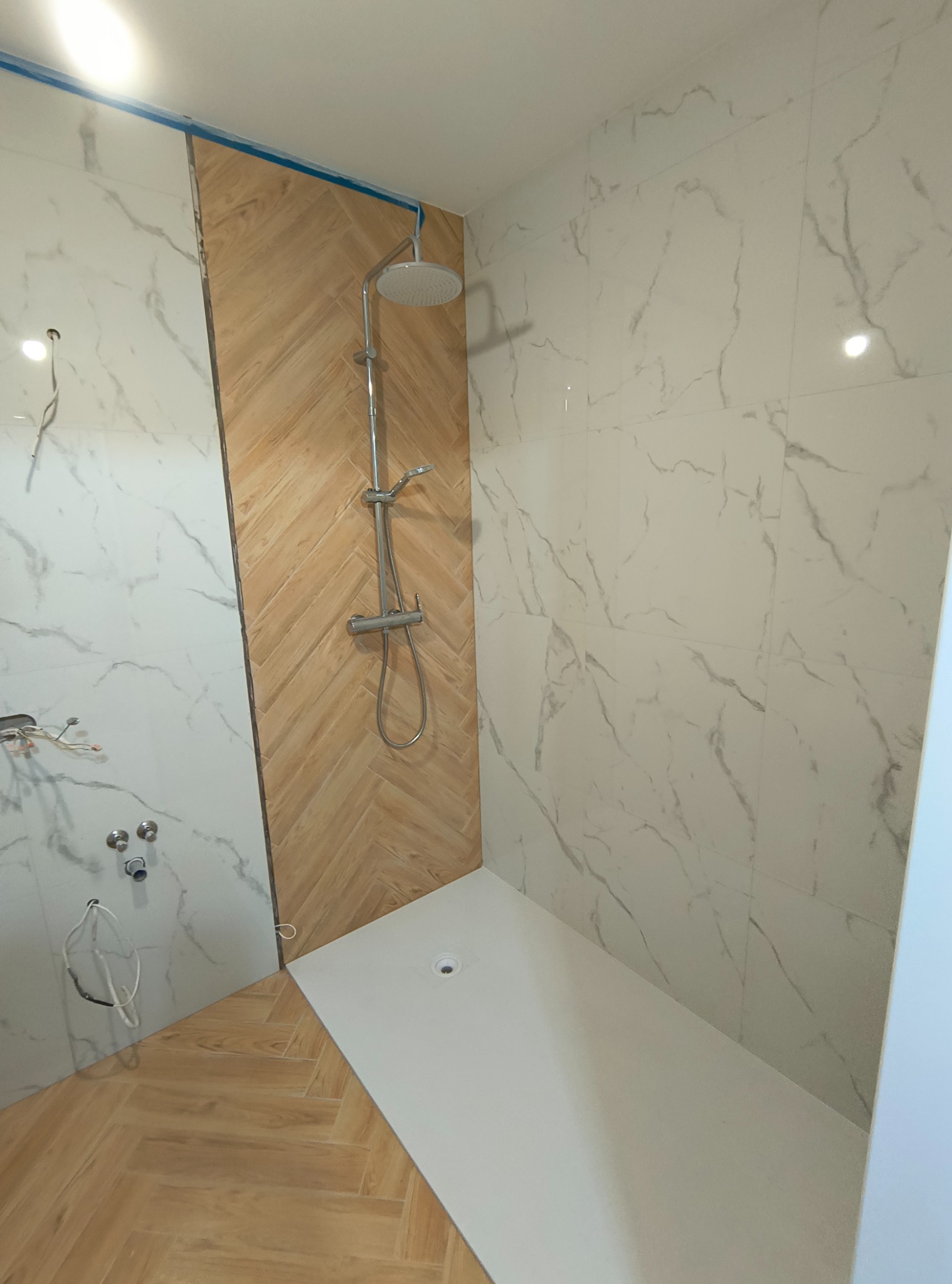 Montaż kabiny prysznicowej z marmurowymi i drewnopodobnymi płytkami, widoczne niedokończone podłączenia i zabezpieczenie taśmą malarską