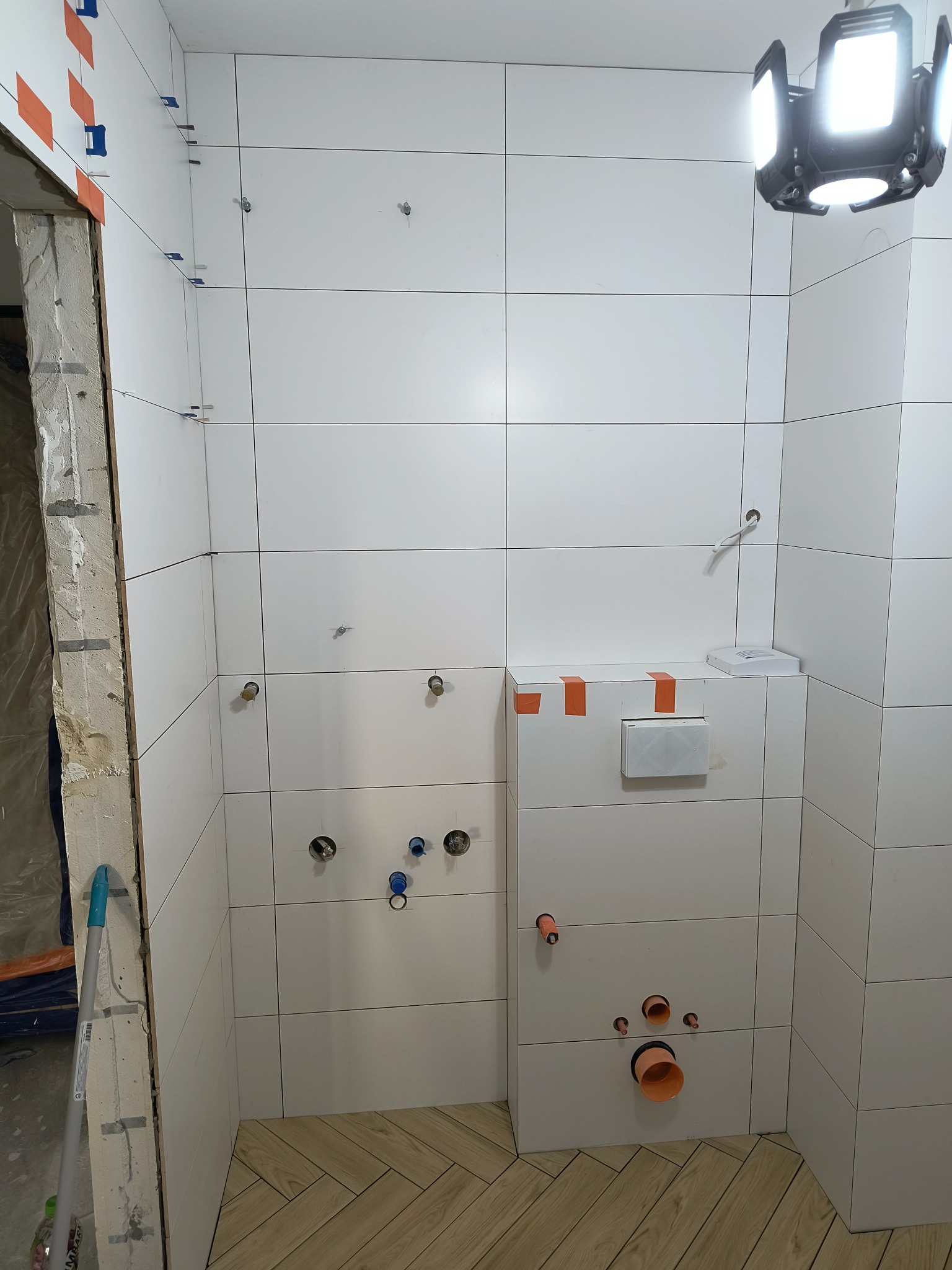 Białe płytki ścienne w łazience w trakcie montażu, widoczne pomarańczowe taśmy i niebieskie kliny poziomujące, otwory na instalacje sanitarne oraz fragment podłogi wyłożonej parkietem w jodełkę.