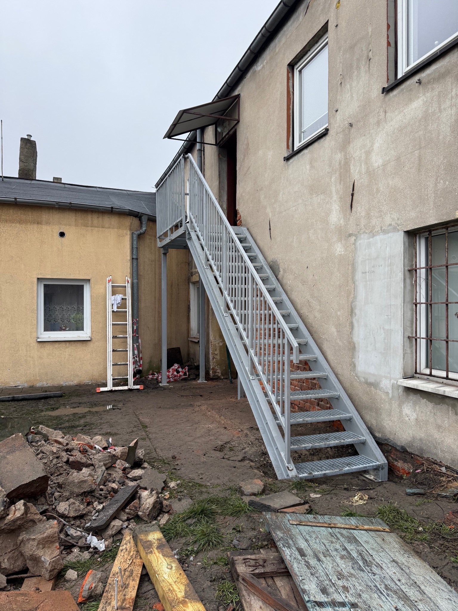 Zewnętrzne schody metalowe z kratowymi stopniami, prowadzące do wejścia w budynku. Obok gruz i drabina oparta o ścianę. Szare niebo.