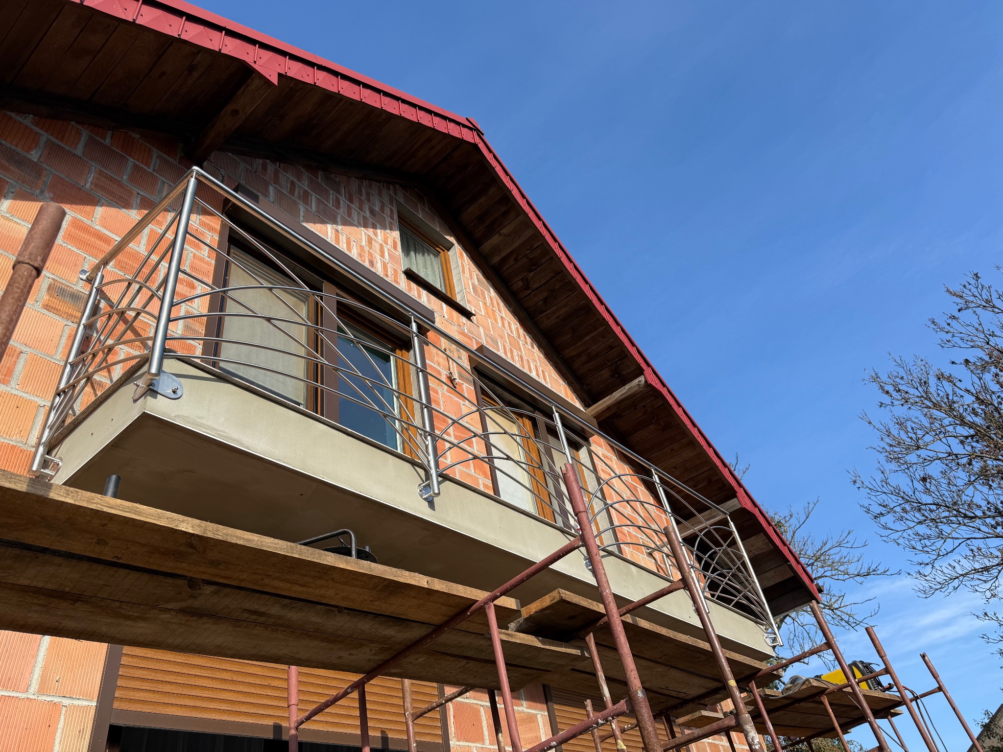 Balkon z czerwoną cegłą i metalową balustradą, widziany z dołu, z rusztowaniem w kadrze. Błękitne niebo w tle. Widoczne okna z brązowymi ramami.