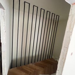 Mateusz Raniewicz - Surowe wnętrze w trakcie wykańczania z widocznymi pionowymi, czarnymi, metalowymi elementami balustrady schodowej, zamontowanymi na białej ścianie, z fragmentem parkietu na podłodze i folią ochronną.