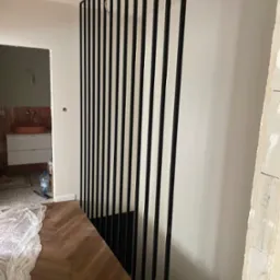 Częściowo zamontowana, czarna, metalowa ścianka działowa z pionowych profili, widoczna na tle remontowanego wnętrza z drewnianą podłogą w jodełkę i elementami wykończenia łazienki.