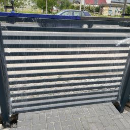 Mateusz Raniewicz - Nowa, pozioma brama metalowa w folii ochronnej, częściowo zamontowana na kostce brukowej, z widoczną niebieską plandeką na górze i samochodem w tle.