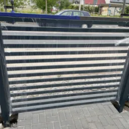 Nowa, pozioma brama metalowa w folii ochronnej, częściowo zamontowana na kostce brukowej, z widoczną niebieską plandeką na górze i samochodem w tle.