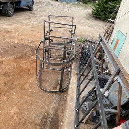 Mateusz Raniewicz - Surowe, metalowe konstrukcje schodowe i poręcze na placu budowy, widoczny niebieski samochód dostawczy i inne elementy metalowe w tle.