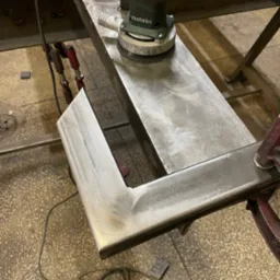 Szlifowanie metalowego profilu w kształcie litery U szlifierką oscylacyjną marki Metabo, zamocowanego klamrami do metalowej konstrukcji stołu warsztatowego.