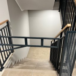 Mateusz Raniewicz - Klatka schodowa z metalowymi schodami i zabezpieczeniem w postaci barierki z drewnianymi poręczami, widok z góry na beżowe płytki schodowe i proste, nowoczesne wykończenie.