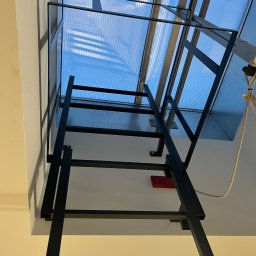 Mateusz Raniewicz - Czarna, metalowa drabina przymocowana do ściany i sufitu, prowadząca do świetlika z poliwęglanu. Widoczne mocowania drabiny do ściany i sufitu, oraz okablowanie elektryczne w pobliżu świetlika.