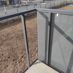Mateusz Raniewicz - Szklana balustrada balkonowa z metalowymi, grafitowymi słupkami, widok z góry na plac budowy z zaparkowanymi samochodami w tle.