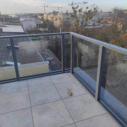 Mateusz Raniewicz - Szklana balustrada balkonowa z aluminiowymi ramami, widok na miasto w tle, lekko pochmurne niebo.
