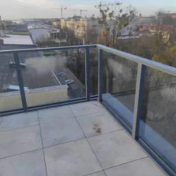 Szklana balustrada balkonowa z aluminiowymi ramami, widok na miasto w tle, lekko pochmurne niebo.