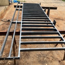 Mateusz Raniewicz - Surowa, metalowa konstrukcja kratowa z profili, wsparta na kozłach spawalniczych na zewnątrz, widoczne ślady spawania i przygotowania do dalszej obróbki.