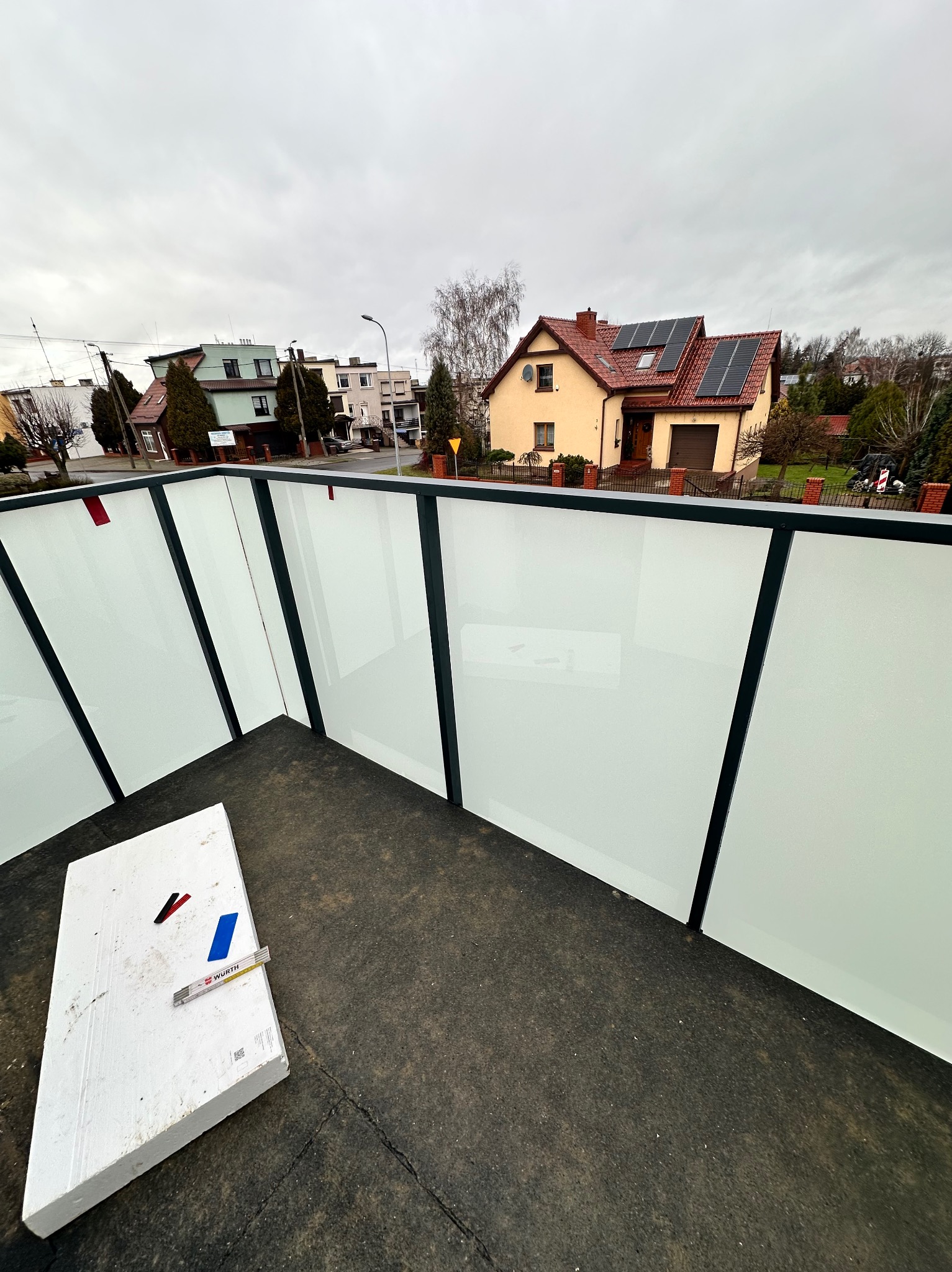 Barierka balkonowa z mlecznego szkła i czarnych metalowych ram, widok z góry na fragment balkonu z leżącą białą płytą styropianową i miarką Wurth, w tle zabudowa jednorodzinna przy pochmurnym niebie.