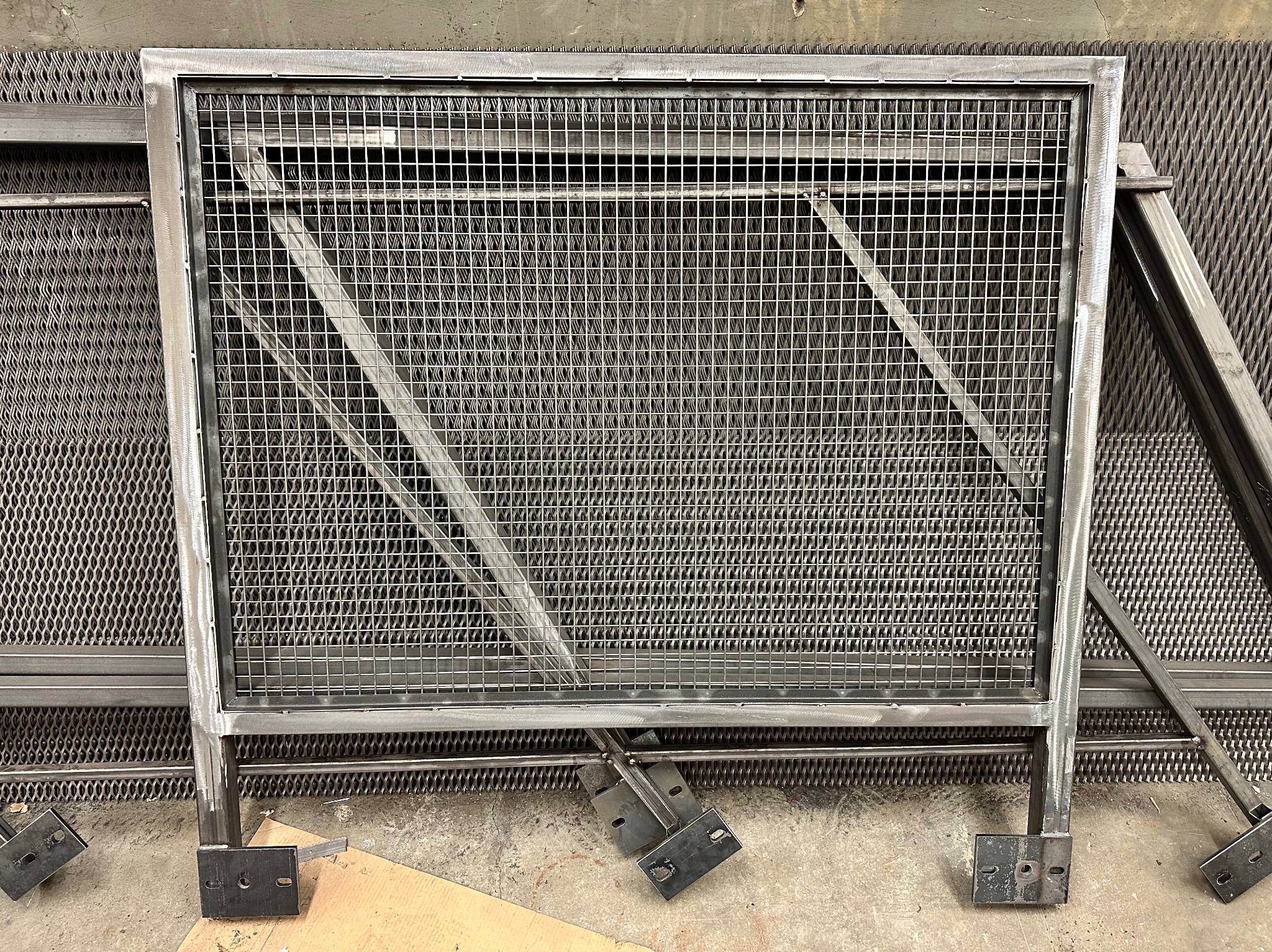 Surowa, metalowa konstrukcja kraty z kwadratową siatką, wsparta na metalowych nogach z widocznymi spawami, oparta o perforowaną blachę w warsztacie.