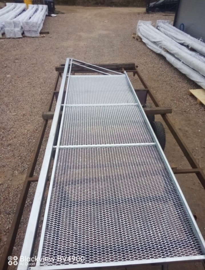 Siatkowa brama wjazdowa z metalową ramą, przewożona na przyczepce, przygotowana do montażu, z widocznymi elementami ogrodzenia w tle.