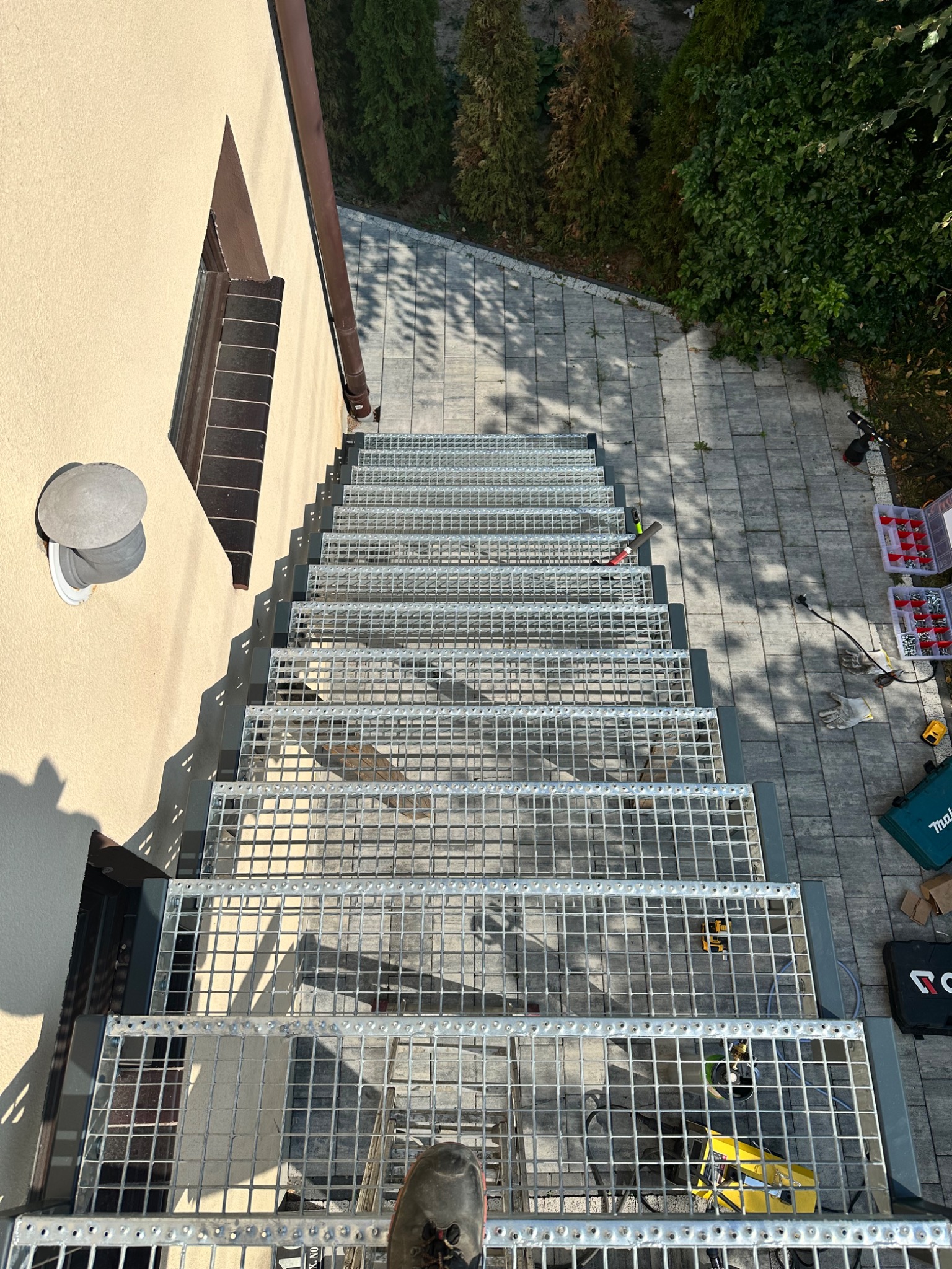 Zewnętrzne metalowe schody kratowe prowadzące z poziomu gruntu do okna na piętrze budynku, widok z góry, częściowo widoczne narzędzia montażowe.