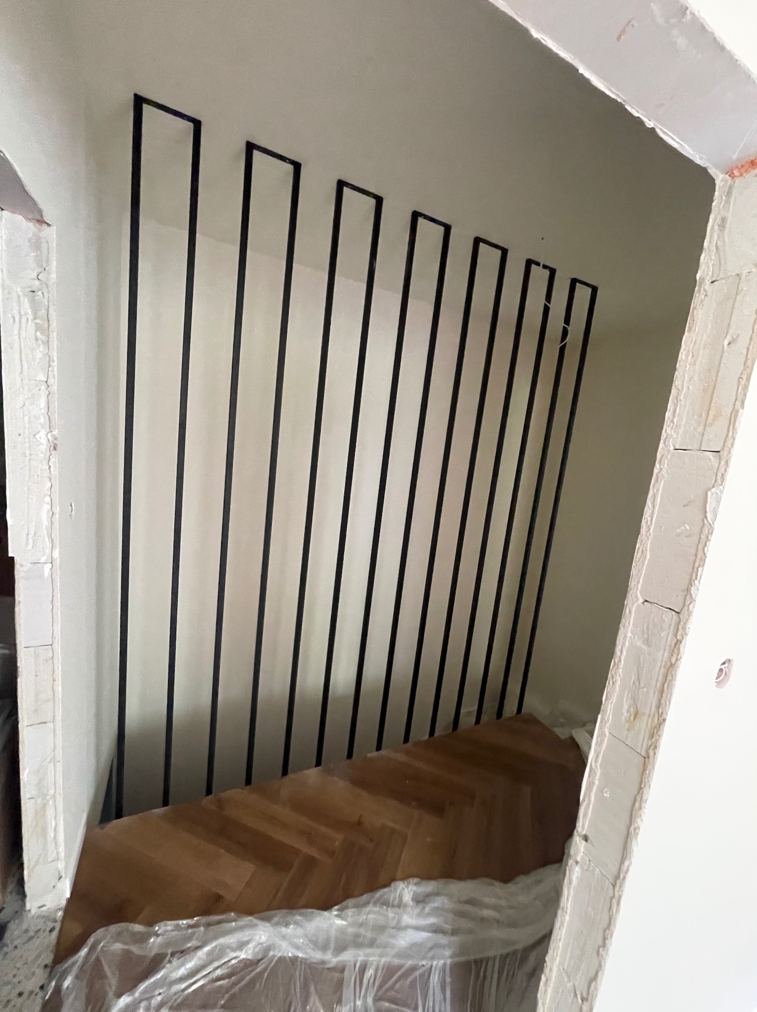 Surowe wnętrze w trakcie wykańczania z widocznymi pionowymi, czarnymi, metalowymi elementami balustrady schodowej, zamontowanymi na białej ścianie, z fragmentem parkietu na podłodze i folią ochronną.
